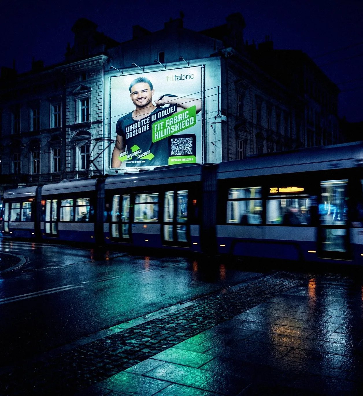 Billboard kampanii otwarcia FitFabric na kamienicy przy torach tramwajowych nocą
